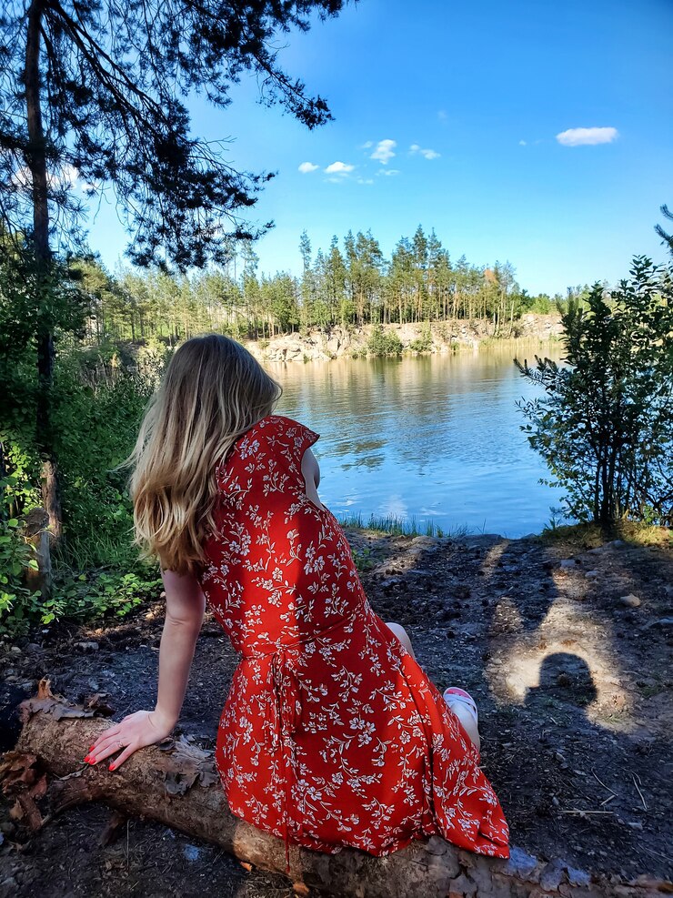 Foto loira com um vestido vermelho, sentado em uma árvore caída na margem de um lago da floresta em tempo ensolarado. resto do campo, natureza, vista traseira