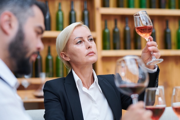 Loira confiante sommelier feminina ou cavista olhando para a cor de um vinho em um copo de vinho no trabalho na adega