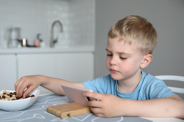 Foto loira de menino bonitinho olha para o smartphone sentado de aveia na mesa em casa. educação on-line do conceito, aprendizagem, jogos.