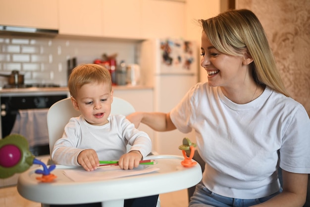 Loira de menino pequeno lindo bebê sentado na cadeira de bebê aprendendo a desenhar a bela jovem mãe e