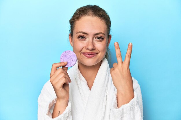 Loira de roupão com esponja facial em estúdio mostrando o número dois com os dedos