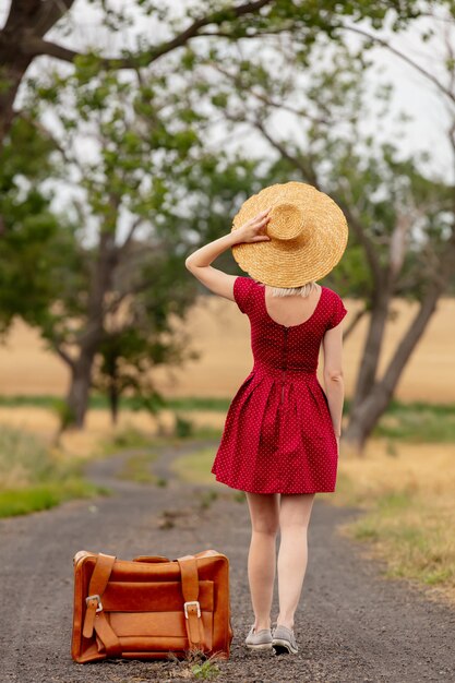 Foto loira de vestido vermelho com uma mala em uma estrada rural antes da chuva