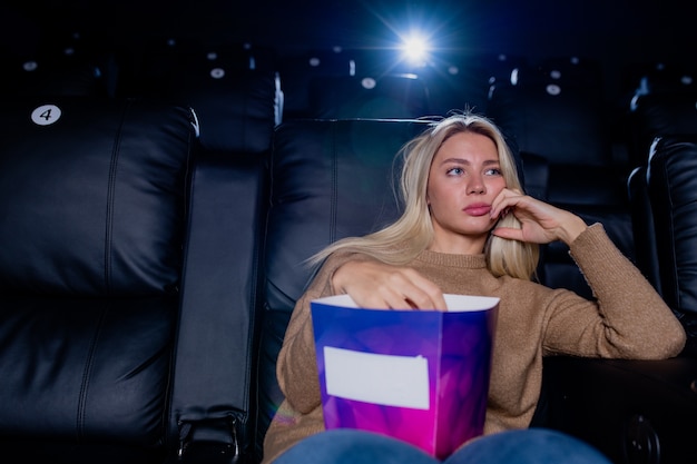 Loira entediada com uma caixa de pipoca sentada na poltrona de couro preto na frente de uma tela grande no cinema e assistindo filme