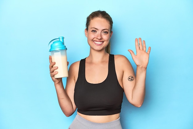 Loira esportiva com um shake de proteína em um estúdio sorrindo alegremente mostrando o número cinco com os dedos