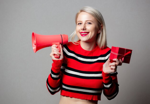 Loira estilizada em suéter vermelho com megafone e caixa de presente na parede cinza