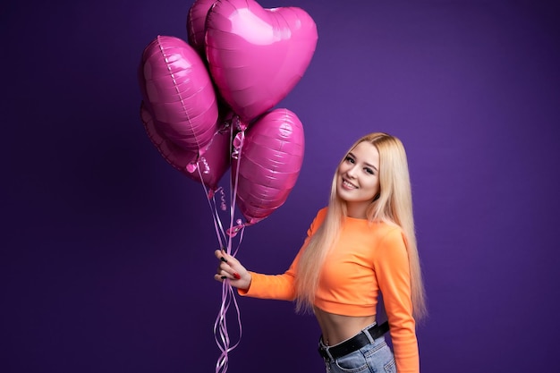 Foto loira feliz com balões em forma de coração em um fundo roxo no estúdio. dia dos namorados.