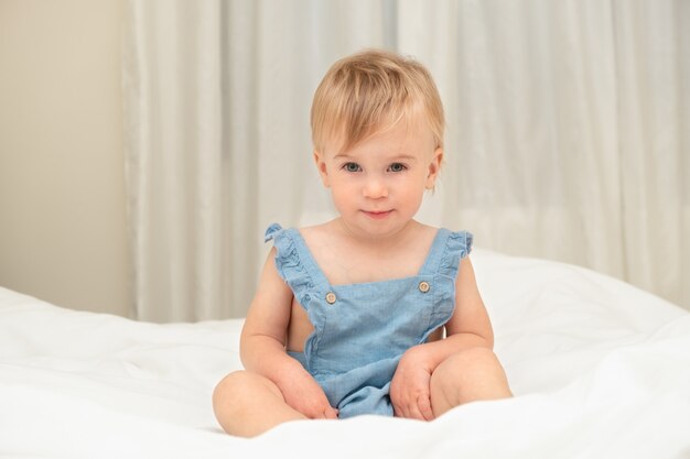Loira fofa, loira, bebê, caucasiana, olhando para a câmera sentada na cama com um cobertor branco em casa