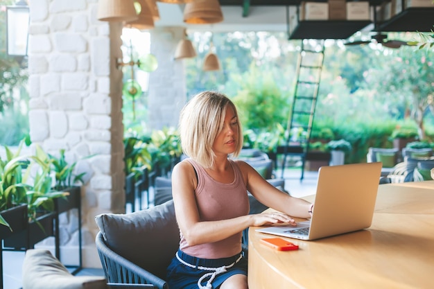 Loira freelancer sentada no ensolarado café ao ar livre, ela está trabalhando usando o laptop. Local de trabalho de escritório aconchegante, trabalho remoto e conceito de trabalho em casa.