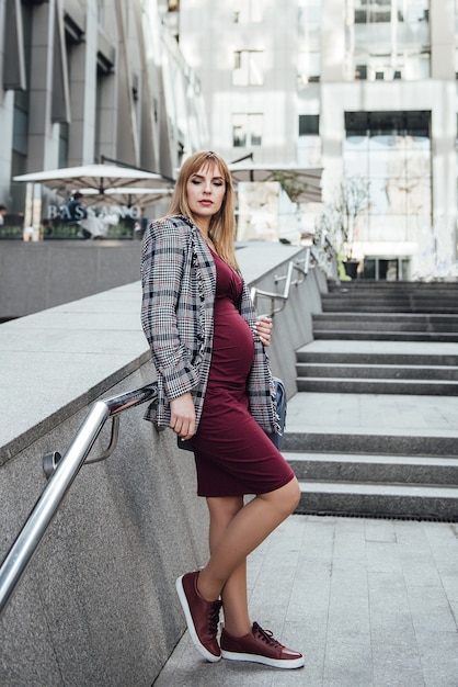 Loira garota grávida em um vestido cor de vinho em forma e uma jaqueta cinza. na cidade grande. danos ao meio ambiente. cabelo longo. 9 meses de antecedência. arranha-céus. urbanismo. felicidade de ser mãe.