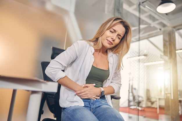 Foto loira infeliz com dor de estômago enquanto está sentada na cadeira do trabalho