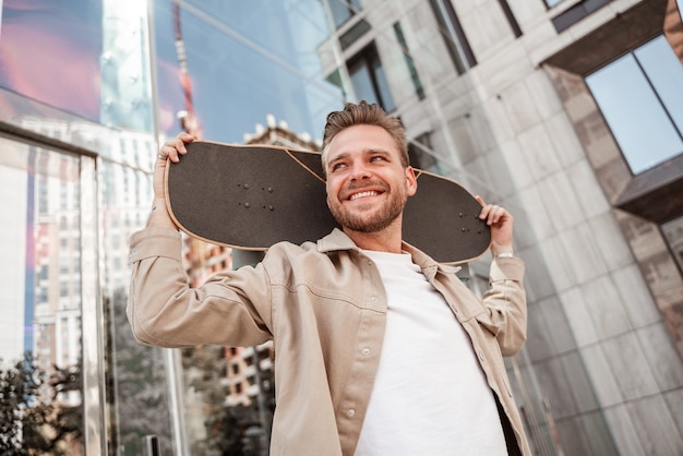 Loira jovem bonita sorrindo homem segurando o skate nos ombros em pé de rua da cidade entre edifícios de vidro. Usa roupa jeans. Skatista esportivo e estiloso pensando em truques extremos lá fora.