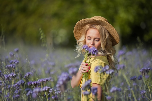 Loira jovem e bonita com um chapéu percorre um campo de flores roxas. Verão. Primavera.