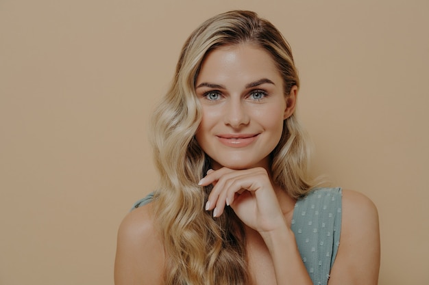 Loira jovem e relaxada linda em um vestido azul com a mão sob o queixo olhando para a câmera com otimismo, satisfação e um leve sorriso, isolado ao lado da parede laranja