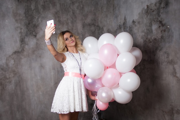Loira jovem encantadora em um vestido branco com balões rosa, faça uma selfie