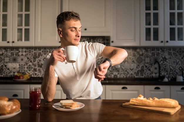 Loira jovem segurando a xícara de café e olhando no relógio na cozinha