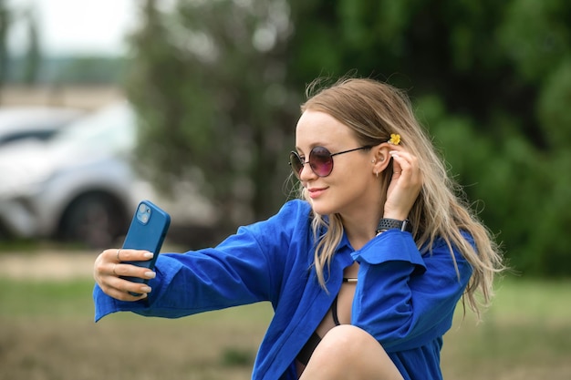 Loira linda com cabelo comprido em uma camisa azul em uma caminhada no parque faz uma selfie no telefone