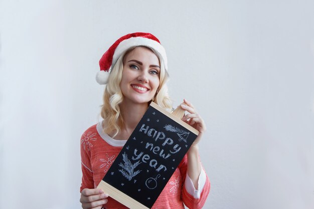 Loira linda com um chapéu de Natal segurando uma placa com a inscrição feliz ano novo e sorrindo