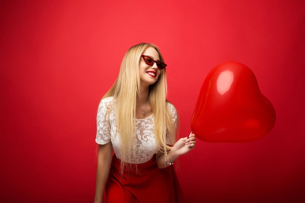 Loira linda e positiva com um balão em forma de coração em um fundo vermelho isolado.