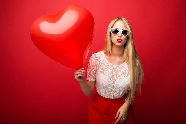 Foto loira linda e positiva com um balão em forma de coração em um fundo vermelho isolado.