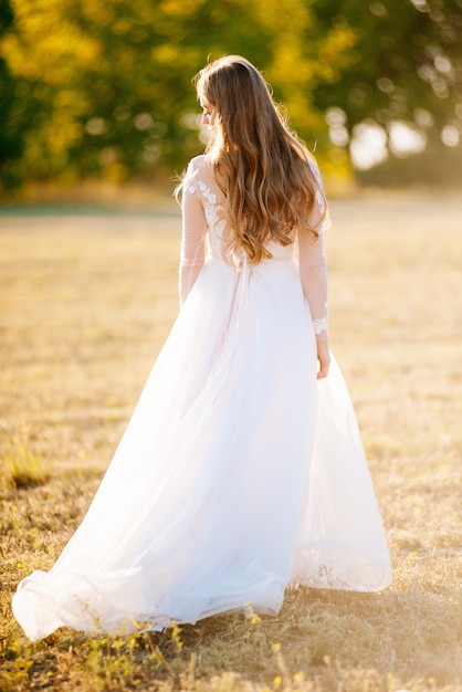 Loira linda e romântica, vestida com um vestido de noiva está andando em um campo, olhando para trás na luz do sol.