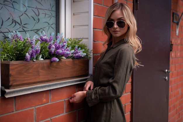 Loira linda em óculos elegantes ajeita o cabelo na janela de uma casa decorada com flores da rua.