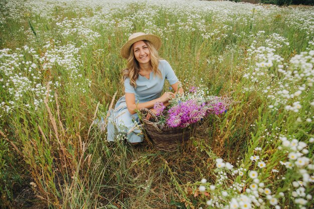 loira linda em um campo de margaridas. mulher em um vestido azul em um campo de flores brancas. menina com um buquê de margaridas. foto concurso de verão na vila. flores silvestres. garota usando um chapéu de palha