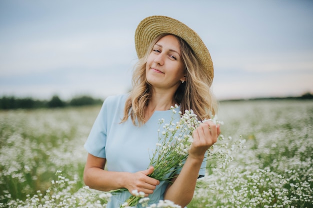 loira linda em um campo de margaridas. mulher em um vestido azul em um campo de flores brancas. menina com um buquê de margaridas. foto concurso de verão na vila. flores silvestres. garota usando um chapéu de palha