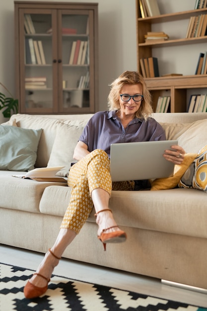 Loira madura e relaxada empresária olhando para a tela do tablet enquanto está sentada no sofá e assistindo a um filme online em casa