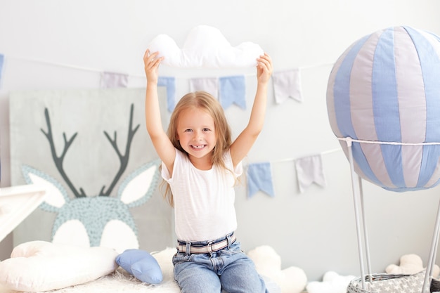 Loira menina sorridente segurando um travesseiro de nuvem na parede de um balão decorativo. A criança brinca no quarto das crianças com brinquedos. O conceito de infância, viajar. Criança no jardim de infância