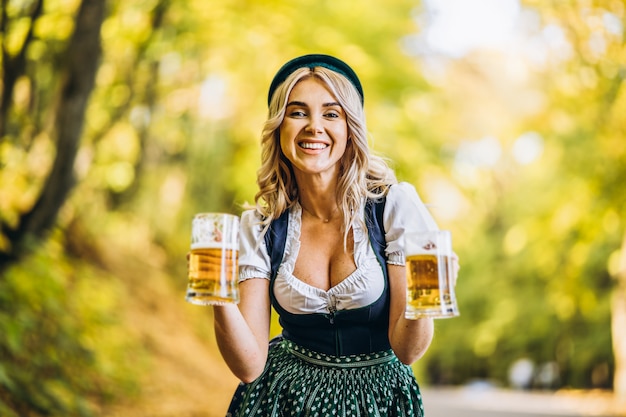 Foto loira muito feliz em vestidos casuais, tradicional festival, segurando duas canecas de cerveja ao ar livre na floresta