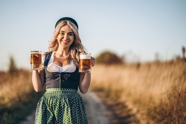 Loira muito feliz em vestidos casuais, tradicional festival, segurando duas canecas de cerveja ao ar livre no campo