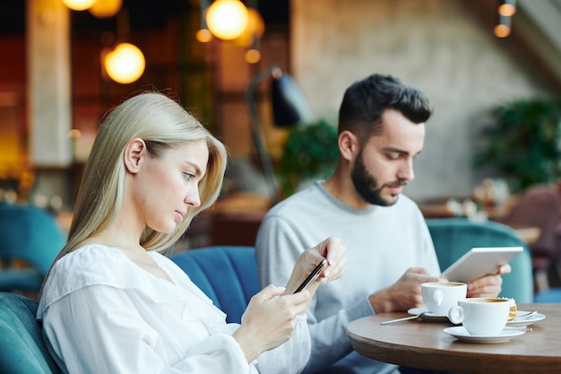 Loira muito séria e o namorado sentados à mesa no café, bebendo e usando dispositivos móveis no lazer