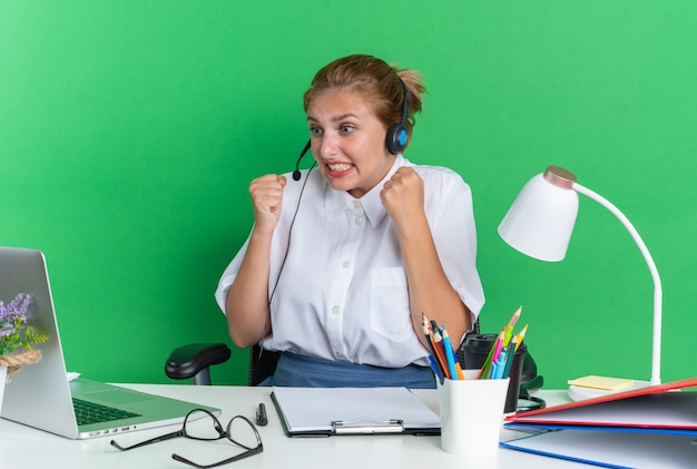 Loira nervosa no call center usando um fone de ouvido, sentada na mesa com as ferramentas de trabalho cerrando os punhos, olhando para o laptop isolado na parede verde