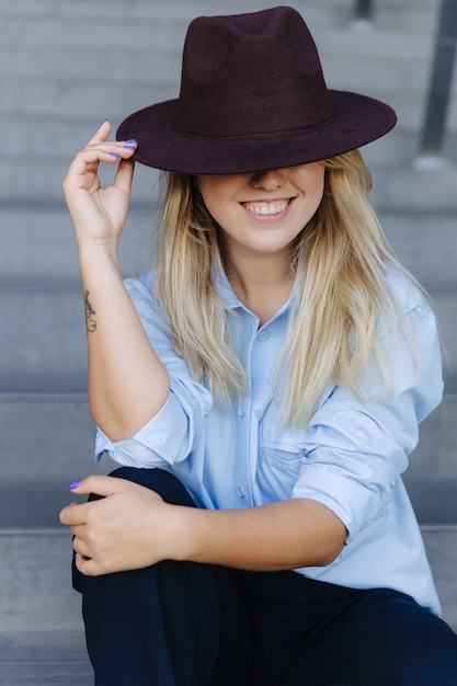 Loira sorridente sentada nos degraus ao ar livre e escondendo os olhos atrás do chapéu da moda. Estilo de vida urbano de hipster feminina. Conceito de estilo.