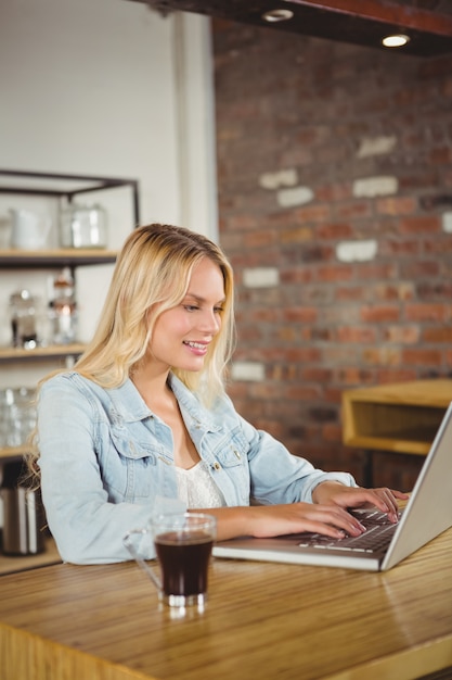 Loira sorridente tomando café e usando o laptop na cafeteria