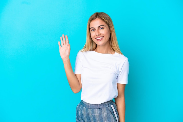 Foto loira uruguaia isolada em fundo azul saudando com a mão com expressão feliz