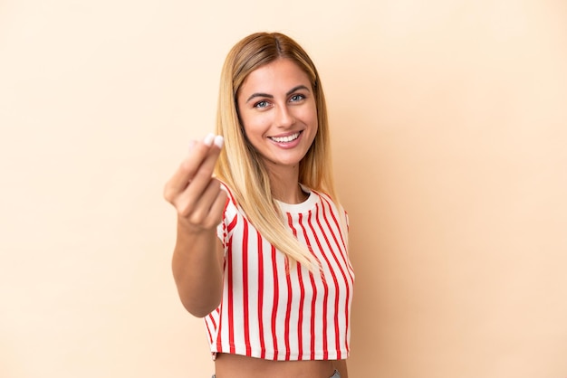 Foto loira uruguaia isolada em fundo bege, fazendo gesto de dinheiro