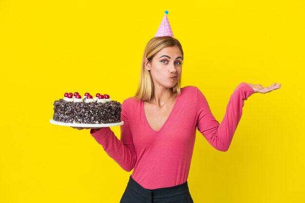 Loira uruguaia segurando bolo de aniversário isolado em fundo amarelo com dúvidas ao levantar as mãos