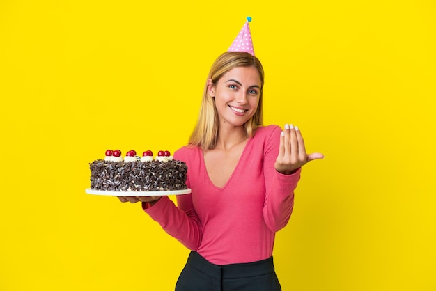 Loira uruguaia segurando bolo de aniversário isolado em fundo amarelo convidando para vir com a mão Feliz que você veio