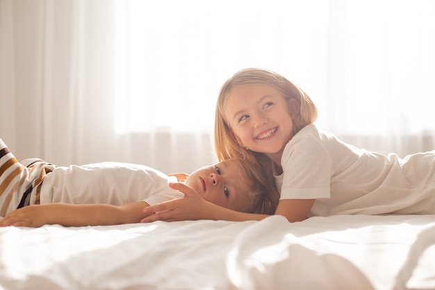 Loiras pequenas, filhos, irmão e irmã em camisetas brancas, estão deitadas na cama em um lençol branco