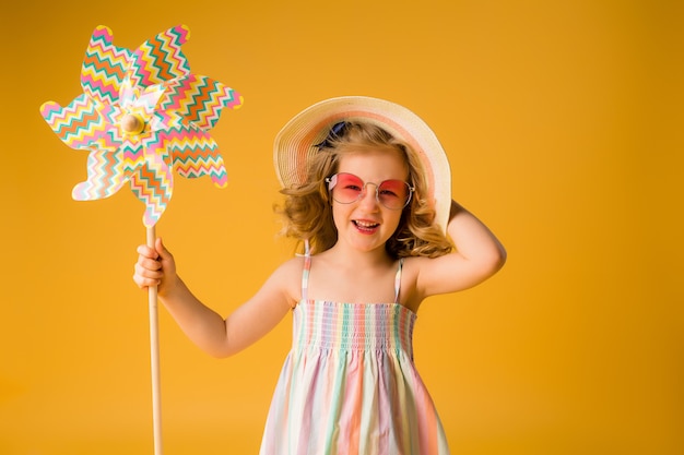 Loirinha sorri em um vestido de verão, óculos de sol, segurando o moinho de vento de uma criança em uma parede amarela