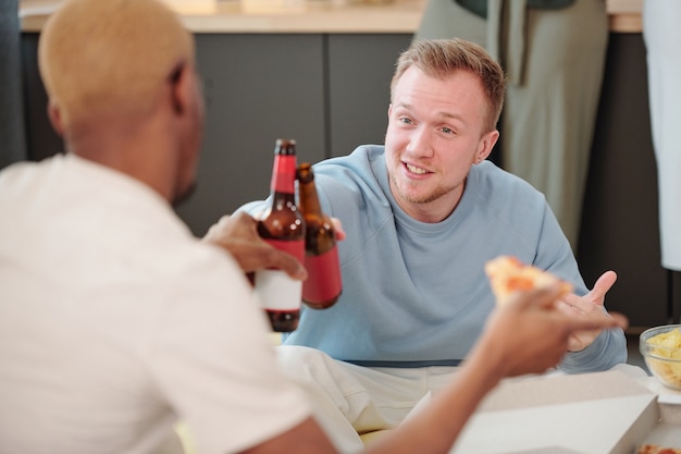 Foto loiro feliz em um moletom azul claro olhando para um jovem africano enquanto os dois tilintam com garrafas de cerveja na mesa e comem lanches