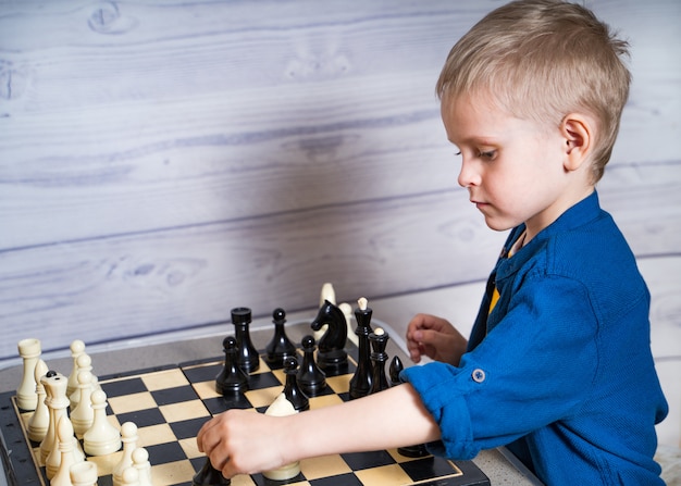 Loiro menino bonitinho está fazendo sua jogada enquanto estiver jogando xadrez. Lógica desenvolvendo jogo de tabuleiro.