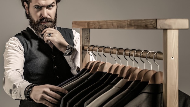 Foto lojas de roupas masculinas alfaiataria terno masculino elegante terno de homem alfaiate em sua oficina bonito homem de moda barbudo em traje clássico ternos masculinos pendurados em uma fileira