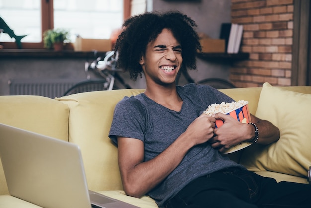 Lol. africano jovem alegre assistindo tv e segurando um balde de pipoca enquanto está sentado no sofá em casa