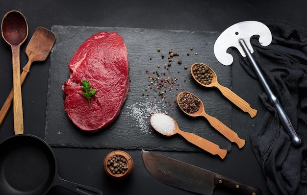 Foto lombo de carne crua encontra-se em uma tábua de corte e especiarias para cozinhar em uma mesa preta