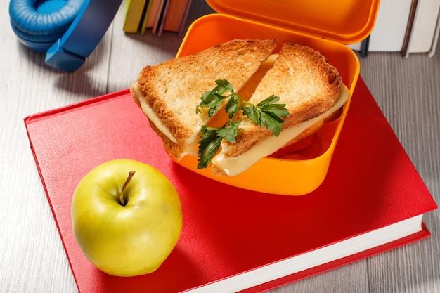 Lonchera amarilla con rebanadas tostadas de pan, queso y perejil verde, manzana verde, libro y auriculares en el fondo. Desayuno escolar.