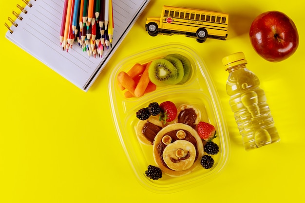 Lonchera para niños con panqueques, bayas, bebida y manzana sobre superficie amarilla