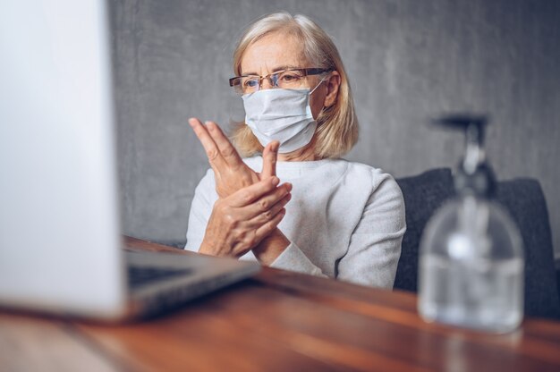 Lonely triste anciana senior en máscara médica facial utilizando desinfectante líquido antibacteriano de mano con computadora portátil en casa cuarentena de auto aislamiento durante la pandemia de coronavirus covid19. quedarse en casa