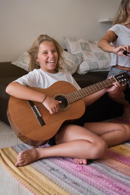 La longitud completa de la chica riendo en ropa casual divirtiéndose mientras está sentada en la alfombra cerca del sofá en la sala de estar y tocando la guitarra acústica el día de fin de semana